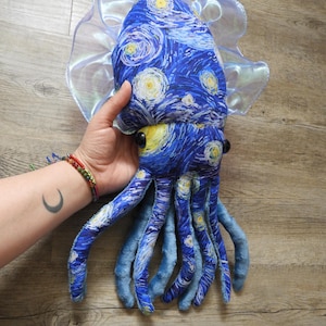 Puede incluir: Un juguete de peluche con forma de calamar con un patr&oacute;n de remolino azul y amarillo que recuerda a "La noche estrellada" de Van Gogh. El juguete tiene grandes ojos negros, tent&aacute;culos azules y un borde transl&uacute;cido y con volantes. El juguete es sostenido por una persona.