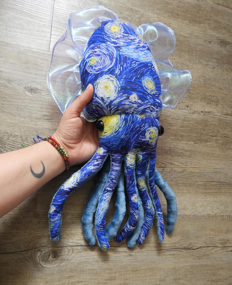 Puede incluir: Un juguete de peluche con forma de calamar con un patr&oacute;n de remolino azul y amarillo que recuerda a "La noche estrellada" de Van Gogh. El juguete tiene grandes ojos negros, tent&aacute;culos azules y un borde transl&uacute;cido y con volantes. El juguete es sostenido por una persona.