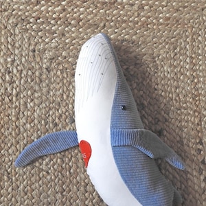 Peut inclure: Une baleine en peluche bleue et blanche, avec un d&eacute;tail en forme de c&oelig;ur rouge. La baleine a une texture c&ocirc;tel&eacute;e et est pos&eacute;e sur une surface beige tiss&eacute;e.