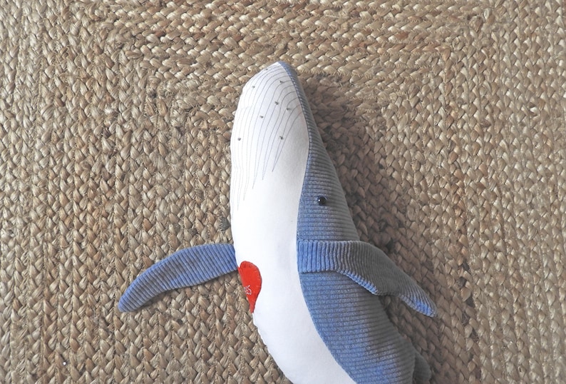 Peut inclure: Une baleine en peluche bleue et blanche, avec un d&eacute;tail en forme de c&oelig;ur rouge. La baleine a une texture c&ocirc;tel&eacute;e et est pos&eacute;e sur une surface beige tiss&eacute;e.