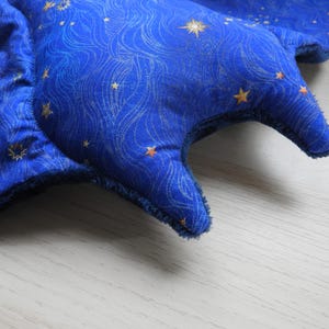 K&ouml;nnte beinhalten: Ein blaues, sternf&ouml;rmiges Pl&uuml;schtier mit einem Himmelsmuster. Der Stoff hat ein Wirbeldesign mit goldenen Sternen. Die R&auml;nder sind mit einem dunkleren blauen Stoff beschnitten. Das Spielzeug liegt auf einer hellen Holzoberfl&auml;che.