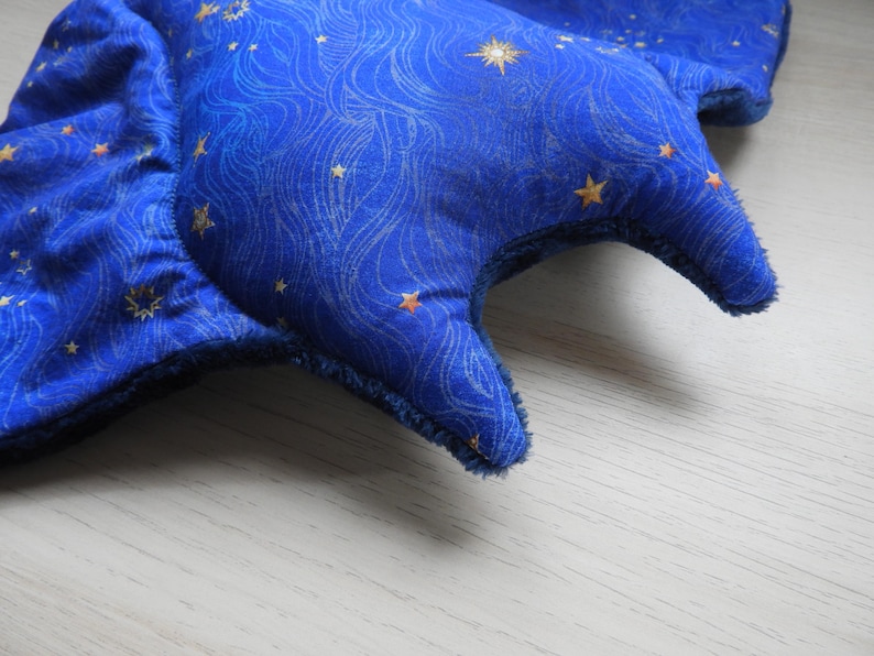 K&ouml;nnte beinhalten: Ein blaues, sternf&ouml;rmiges Pl&uuml;schtier mit einem Himmelsmuster. Der Stoff hat ein Wirbeldesign mit goldenen Sternen. Die R&auml;nder sind mit einem dunkleren blauen Stoff beschnitten. Das Spielzeug liegt auf einer hellen Holzoberfl&auml;che.