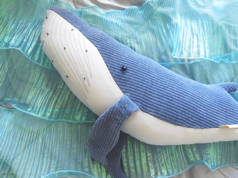 Peut inclure: Une peluche de baleine bleue avec un ventre blanc et une nageoire caudale d&eacute;taill&eacute;e. La baleine est pos&eacute;e sur une surface textur&eacute;e bleue et verte, cr&eacute;ant un effet marin. L'&oelig;il de la baleine est une petite perle noire.