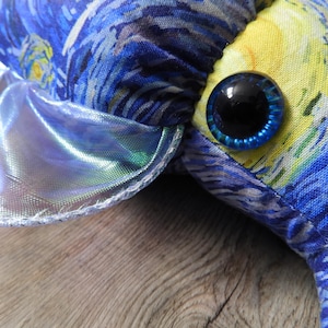 Puede incluir: Primer plano de un juguete de peluche con una tela estampada de noche estrellada azul y amarilla. El juguete tiene un ojo grande, redondo y azul con una pupila negra. Una tela plateada brillante e iridiscente est&aacute; cosida alrededor del borde del juguete.