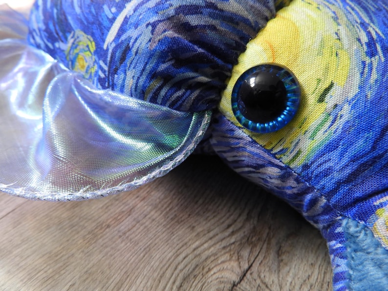 Puede incluir: Primer plano de un juguete de peluche con una tela estampada de noche estrellada azul y amarilla. El juguete tiene un ojo grande, redondo y azul con una pupila negra. Una tela plateada brillante e iridiscente est&aacute; cosida alrededor del borde del juguete.