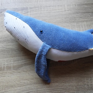 Peut inclure: Une peluche baleine bleue en tissu c&ocirc;tel&eacute;. La baleine a un ventre blanc et un visage blanc avec des d&eacute;tails de points noirs. Le jouet a une petite nageoire et une nageoire caudale. La baleine est pos&eacute;e sur une surface en bois.