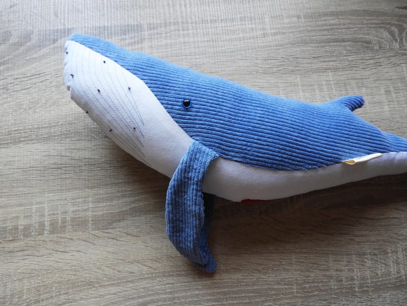 Peut inclure: Une peluche baleine bleue en tissu c&ocirc;tel&eacute;. La baleine a un ventre blanc et un visage blanc avec des d&eacute;tails de points noirs. Le jouet a une petite nageoire et une nageoire caudale. La baleine est pos&eacute;e sur une surface en bois.