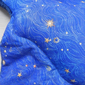 K&ouml;nnte beinhalten: Nahaufnahme eines blauen Stoffes mit einem Himmelsdesign. Der Stoff weist ein Muster aus wirbelnden Linien und goldenen Sternen in verschiedenen Gr&ouml;&szlig;en auf. Ein gro&szlig;er, verzierter Stern ist ebenfalls sichtbar und verleiht dem Design einen Hauch von Eleganz. Der Stoff hat eine weiche, gepolsterte Textur.