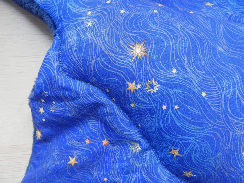 K&ouml;nnte beinhalten: Nahaufnahme eines blauen Stoffes mit einem Himmelsdesign. Der Stoff weist ein Muster aus wirbelnden Linien und goldenen Sternen in verschiedenen Gr&ouml;&szlig;en auf. Ein gro&szlig;er, verzierter Stern ist ebenfalls sichtbar und verleiht dem Design einen Hauch von Eleganz. Der Stoff hat eine weiche, gepolsterte Textur.