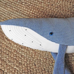 Peut inclure: Une peluche baleine bleue avec un ventre et un visage blancs. La baleine a une texture en velours c&ocirc;tel&eacute; et des d&eacute;tails brod&eacute;s, dont des points noirs et des lignes courbes sur le visage. L'&oelig;il est bleu fonc&eacute;.
