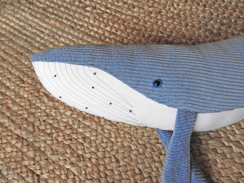 Peut inclure: Une peluche baleine bleue avec un ventre et un visage blancs. La baleine a une texture en velours c&ocirc;tel&eacute; et des d&eacute;tails brod&eacute;s, dont des points noirs et des lignes courbes sur le visage. L'&oelig;il est bleu fonc&eacute;.