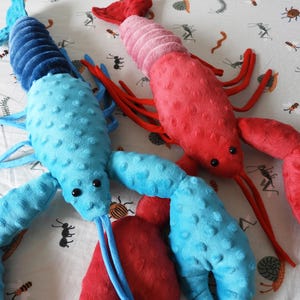 May include: Two plush lobster toys in the image. One is light blue with dark blue accents, and the other is red with pink accents. Both have black eyes and long, thin antennae. The toys are on a white patterned surface.