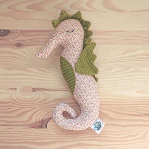 Puede incluir: Un juguete de peluche con forma de caballito de mar con un cuerpo estampado en tonos rosa, blanco y dorado. El caballito de mar tiene aletas de pana verde y una aleta verde en la cabeza. El juguete está sobre una superficie de madera marrón claro.