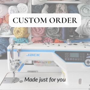 May include: A white and blue Jack A5E sewing machine with the text "CUSTOM ORDER" and "Made just for you". The machine is on a white surface, with shelves of colorful fabrics in the background.