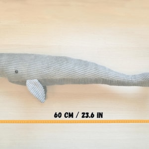 K&ouml;nnte beinhalten: Ein graues Cord-Wal-Pl&uuml;schtier, ca. 60 cm lang, mit kleiner Flosse und Knopfauge. Das Spielzeug liegt auf einer hellen Holzoberfl&auml;che. Der K&ouml;rper des Wals hat eine gerippte Textur.