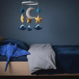 K&ouml;nnte beinhalten: Ein Kinderzimmer mit einem Holzbett und blauer Bettw&auml;sche. Ein Pl&uuml;sch-Stechrochen-Kissen mit Sternenmuster liegt auf dem Bett. Ein Mobile mit Mond, Sternen und Meereskreaturen h&auml;ngt dar&uuml;ber und schafft eine beruhigende Atmosph&auml;re.