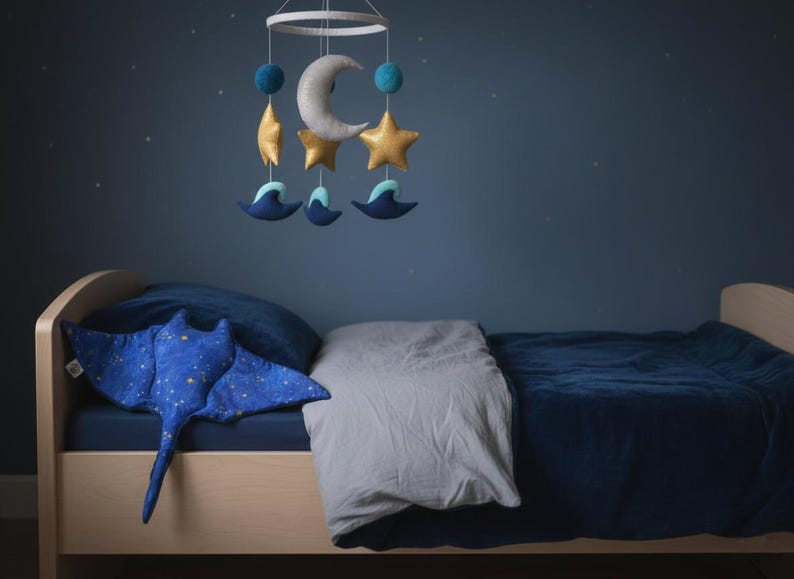 K&ouml;nnte beinhalten: Ein Kinderzimmer mit einem Holzbett und blauer Bettw&auml;sche. Ein Pl&uuml;sch-Stechrochen-Kissen mit Sternenmuster liegt auf dem Bett. Ein Mobile mit Mond, Sternen und Meereskreaturen h&auml;ngt dar&uuml;ber und schafft eine beruhigende Atmosph&auml;re.