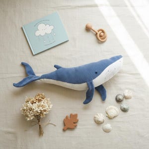 Peut inclure: Une peluche baleine bleue avec un ventre blanc, un hochet en bois, un livre pour enfants, des coquillages et une feuille en bois sur une surface claire. Le livre pr&eacute;sente un motif de nuage et d'&eacute;toile. Des fleurs s&eacute;ch&eacute;es sont &eacute;galement pr&eacute;sentes.