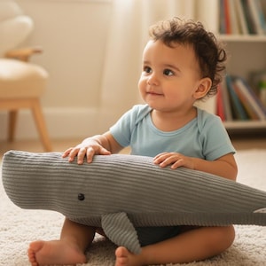 K&ouml;nnte beinhalten: Ein Baby in einem hellblauen Strampler sitzt auf einem cremefarbenen Teppich und h&auml;lt ein graues Cord-Wal-Pl&uuml;schtier. Der Wal hat ein kleines schwarzes Auge und eine Flosse. Das Baby l&auml;chelt und schaut zur Seite.