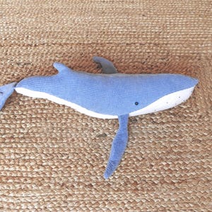 Peut inclure: Une peluche baleine bleue avec un ventre blanc et un petit &oelig;il noir. La baleine a un aileron dorsal et deux nageoires lat&eacute;rales. Le jouet est pos&eacute; sur une surface brune tiss&eacute;e et textur&eacute;e.