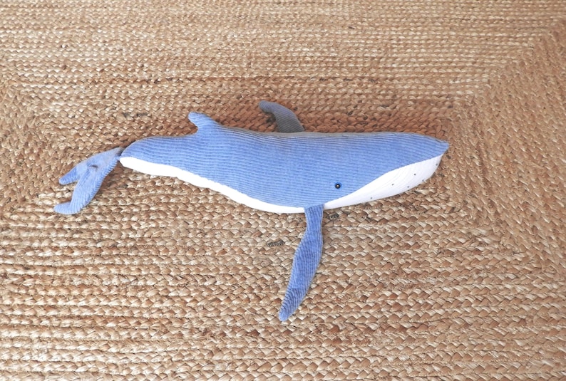 Peut inclure: Une peluche baleine bleue avec un ventre blanc et un petit &oelig;il noir. La baleine a un aileron dorsal et deux nageoires lat&eacute;rales. Le jouet est pos&eacute; sur une surface brune tiss&eacute;e et textur&eacute;e.