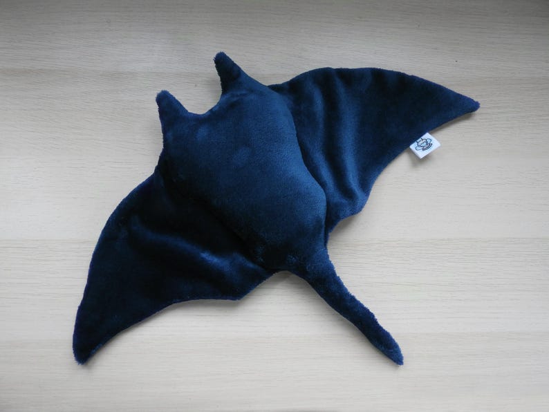K&ouml;nnte beinhalten: Ein dunkelblaues Pl&uuml;sch-Manta-Rochen-Spielzeug mit weicher, samtiger Textur. Das Spielzeug hat fl&uuml;gelartige Flossen und einen langen Schwanz. Ein kleines wei&szlig;es Etikett ist an einer der Flossen befestigt.