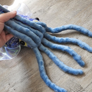 Puede incluir: Un juguete de peluche de calamar azul con largos tent&aacute;culos. El juguete tiene ojos amarillos grandes y una cabeza estampada. El calamar se sostiene en una mano, con un suelo de madera en el fondo.