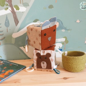 Può includere: Blocchi di tessuto impilabili per bambini con un orso marrone, un animale maculato marrone e bianco e un viso di orso marrone su ogni lato. I blocchi sono su una superficie di legno con un libro e un cesto intrecciato verde.