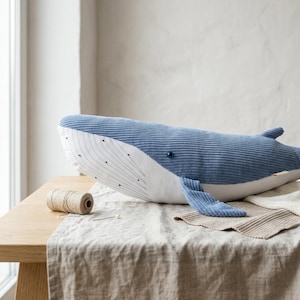 Peut inclure: Une grande baleine en peluche bleue avec une texture en velours c&ocirc;tel&eacute;. La baleine a un ventre blanc et un petit &oelig;il bleu fonc&eacute;. Elle repose sur une nappe en lin beige sur une table en bois. Une bobine de ficelle et une aiguille sont &eacute;galement sur la table.