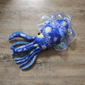 Puede incluir: Un juguete de peluche de pulpo con un patr&oacute;n de noche estrellada azul y amarillo en la cabeza y los tent&aacute;culos. El juguete tiene un cuello transparente y con volantes y un ojo amarillo. El juguete est&aacute; sobre una superficie de madera.