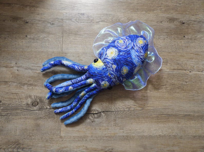 Puede incluir: Un juguete de peluche de pulpo con un patr&oacute;n de noche estrellada azul y amarillo en la cabeza y los tent&aacute;culos. El juguete tiene un cuello transparente y con volantes y un ojo amarillo. El juguete est&aacute; sobre una superficie de madera.