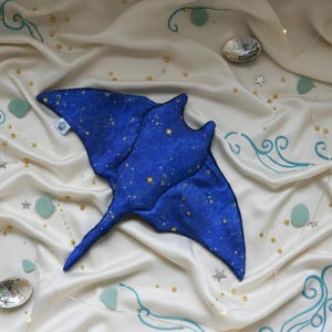 K&ouml;nnte beinhalten: Ein blaues Stofftier in Form eines Mantarochens mit Sternenmuster, das auf cremefarbenem Satin liegt. Der Stoff ist mit t&uuml;rkisfarbenen Wellenmustern, kleinen Sternen und goldenen Perlen verziert. Zwei schillernde Muscheln sind ebenfalls vorhanden.