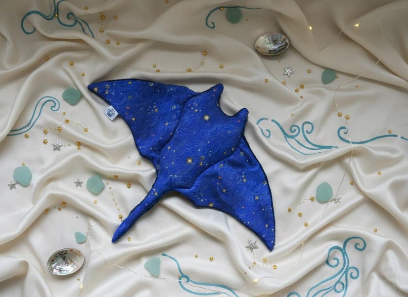 K&ouml;nnte beinhalten: Ein blaues Stofftier in Form eines Mantarochens mit Sternenmuster, das auf cremefarbenem Satin liegt. Der Stoff ist mit t&uuml;rkisfarbenen Wellenmustern, kleinen Sternen und goldenen Perlen verziert. Zwei schillernde Muscheln sind ebenfalls vorhanden.
