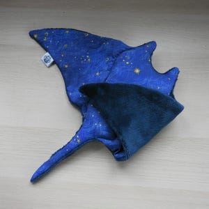 K&ouml;nnte beinhalten: Ein blaues Stoffspielzeug in Form eines Mantarochens mit Sternenhimmelmuster. Das Spielzeug hat eine dunkelblaue Unterseite und ein kleines wei&szlig;es Etikett. Die Fl&uuml;gel des Mantarochens sind ausgebreitet.