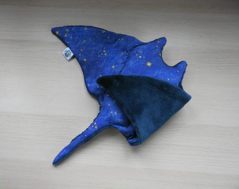 K&ouml;nnte beinhalten: Ein blaues Stoffspielzeug in Form eines Mantarochens mit Sternenhimmelmuster. Das Spielzeug hat eine dunkelblaue Unterseite und ein kleines wei&szlig;es Etikett. Die Fl&uuml;gel des Mantarochens sind ausgebreitet.