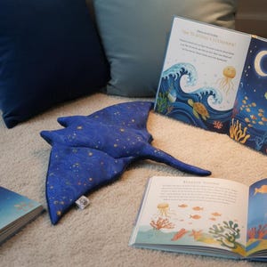K&ouml;nnte beinhalten: Ein blaues Pl&uuml;sch-Stechrochen mit goldenen Sternen liegt auf einem hellen Teppich, umgeben von offenen Kinderb&uuml;chern mit Illustrationen von Meeresbewohnern. Dunkelblaue Kissen bilden den Hintergrund.