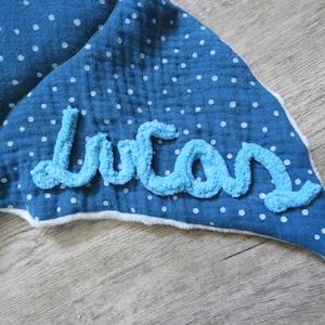 K&ouml;nnte beinhalten: Eine blaue Babydecke mit wei&szlig;en Punkten und dem Namen "Lucas" in hellblauem, flauschigem Garn gestickt. Die Decke hat eine wei&szlig;e Bord&uuml;re und liegt auf einer Holzoberfl&auml;che.