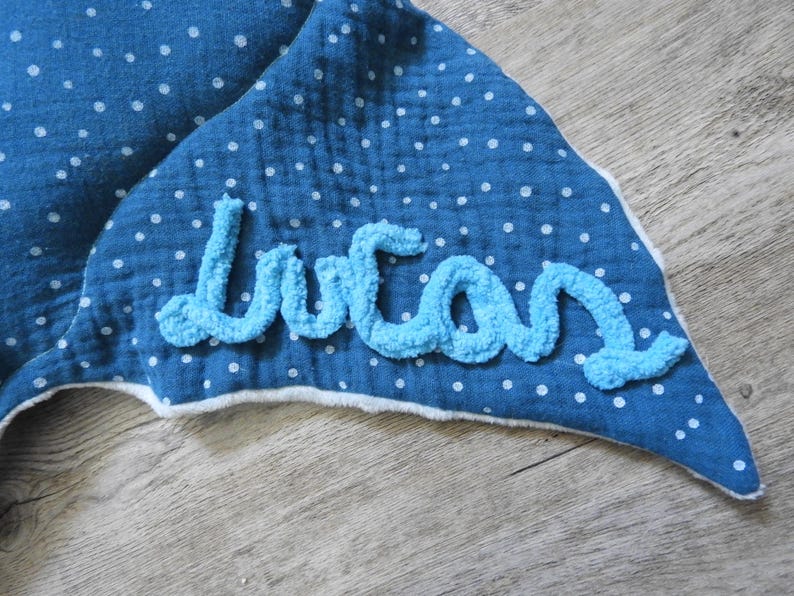 K&ouml;nnte beinhalten: Eine blaue Babydecke mit wei&szlig;en Punkten und dem Namen "Lucas" in hellblauem, flauschigem Garn gestickt. Die Decke hat eine wei&szlig;e Bord&uuml;re und liegt auf einer Holzoberfl&auml;che.