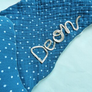 K&ouml;nnte beinhalten: Ein blaues, sternf&ouml;rmiges Baby-Accessoire mit wei&szlig;en Tupfen. Der Name "Deon" ist mit silbernem Faden auf der Ecke gestickt. Der Stoff hat ein strukturiertes, geknittertes Aussehen.