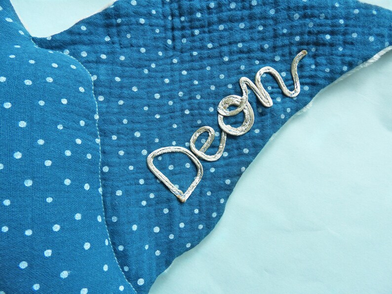 K&ouml;nnte beinhalten: Ein blaues, sternf&ouml;rmiges Baby-Accessoire mit wei&szlig;en Tupfen. Der Name "Deon" ist mit silbernem Faden auf der Ecke gestickt. Der Stoff hat ein strukturiertes, geknittertes Aussehen.