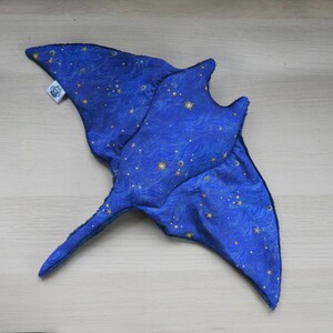 K&ouml;nnte beinhalten: Ein blaues Stoffspielzeug in Form eines Mantarochens mit einem Himmelsmuster aus goldenen Sternen und wellenf&ouml;rmigen Linien. Das Spielzeug hat einen langen Schwanz und fl&uuml;gelf&ouml;rmige Flossen. Ein kleines wei&szlig;es Etikett ist angebracht.