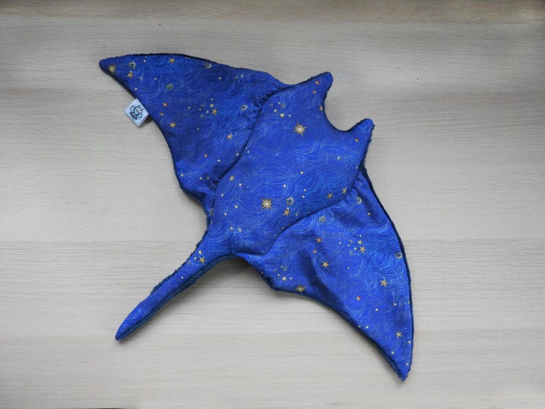 K&ouml;nnte beinhalten: Ein blaues Stoffspielzeug in Form eines Mantarochens mit einem Himmelsmuster aus goldenen Sternen und wellenf&ouml;rmigen Linien. Das Spielzeug hat einen langen Schwanz und fl&uuml;gelf&ouml;rmige Flossen. Ein kleines wei&szlig;es Etikett ist angebracht.