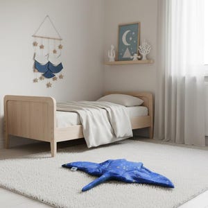 K&ouml;nnte beinhalten: Ein Kinderzimmer mit einem hellen Holzbett und wei&szlig;er Bettw&auml;sche. Ein blaues, rochenf&ouml;rmiges Kissen mit wei&szlig;en Sternendetails liegt auf einem wei&szlig;en Teppich. Ein Wandbehang mit Sternen und ein gerahmtes Bild sind ebenfalls zu sehen.