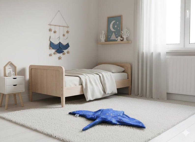 K&ouml;nnte beinhalten: Ein Kinderzimmer mit einem hellen Holzbett und wei&szlig;er Bettw&auml;sche. Ein blaues, rochenf&ouml;rmiges Kissen mit wei&szlig;en Sternendetails liegt auf einem wei&szlig;en Teppich. Ein Wandbehang mit Sternen und ein gerahmtes Bild sind ebenfalls zu sehen.