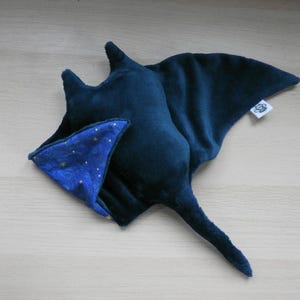 K&ouml;nnte beinhalten: Ein Pl&uuml;schtier in Form eines marineblauen Mantarochens mit einem kontrastierenden blau-goldenen Sternenmuster auf dem Bauch. Das Spielzeug hat einen langen Schwanz und ein kleines Etikett. Das Spielzeug ist aus weichem Stoff.