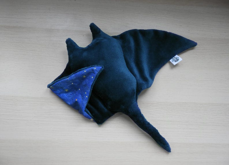 K&ouml;nnte beinhalten: Ein Pl&uuml;schtier in Form eines marineblauen Mantarochens mit einem kontrastierenden blau-goldenen Sternenmuster auf dem Bauch. Das Spielzeug hat einen langen Schwanz und ein kleines Etikett. Das Spielzeug ist aus weichem Stoff.
