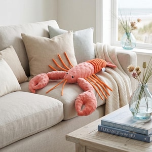 Pode incluir: Um grande brinquedo de pelúcia em forma de lagosta, em tons de coral e laranja, repousa sobre um sofá bege. O brinquedo tem garras detalhadas e longas antenas. O sofá é adornado com almofadas em tons neutros e uma manta creme.