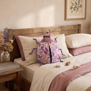 May include: A decorative throw cushion shaped like a beetle, featuring a purple head and six legs. The body is covered in a floral pattern with shades of purple, orange, and white. The cushion is positioned on a bed with cream and mauve bedding.