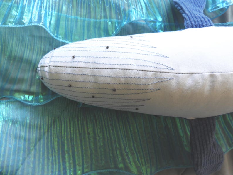 Peut inclure: Un jouet en peluche en forme de baleine blanche avec des d&eacute;tails cousus en bleu et de petits accents noirs. La baleine est plac&eacute;e sur un fond de tissu turquoise chatoyant, cr&eacute;ant un th&egrave;me aquatique et fantaisiste.
