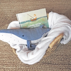 Peut inclure: Un panier en osier contient une peluche baleine bleue, un livre pour enfants et une couverture blanche. La couverture du livre pr&eacute;sente une illustration d'une personne p&ecirc;chant depuis un bateau. Le texte du livre est "&iquest;D&oacute;nde est&aacute;n los peces?"